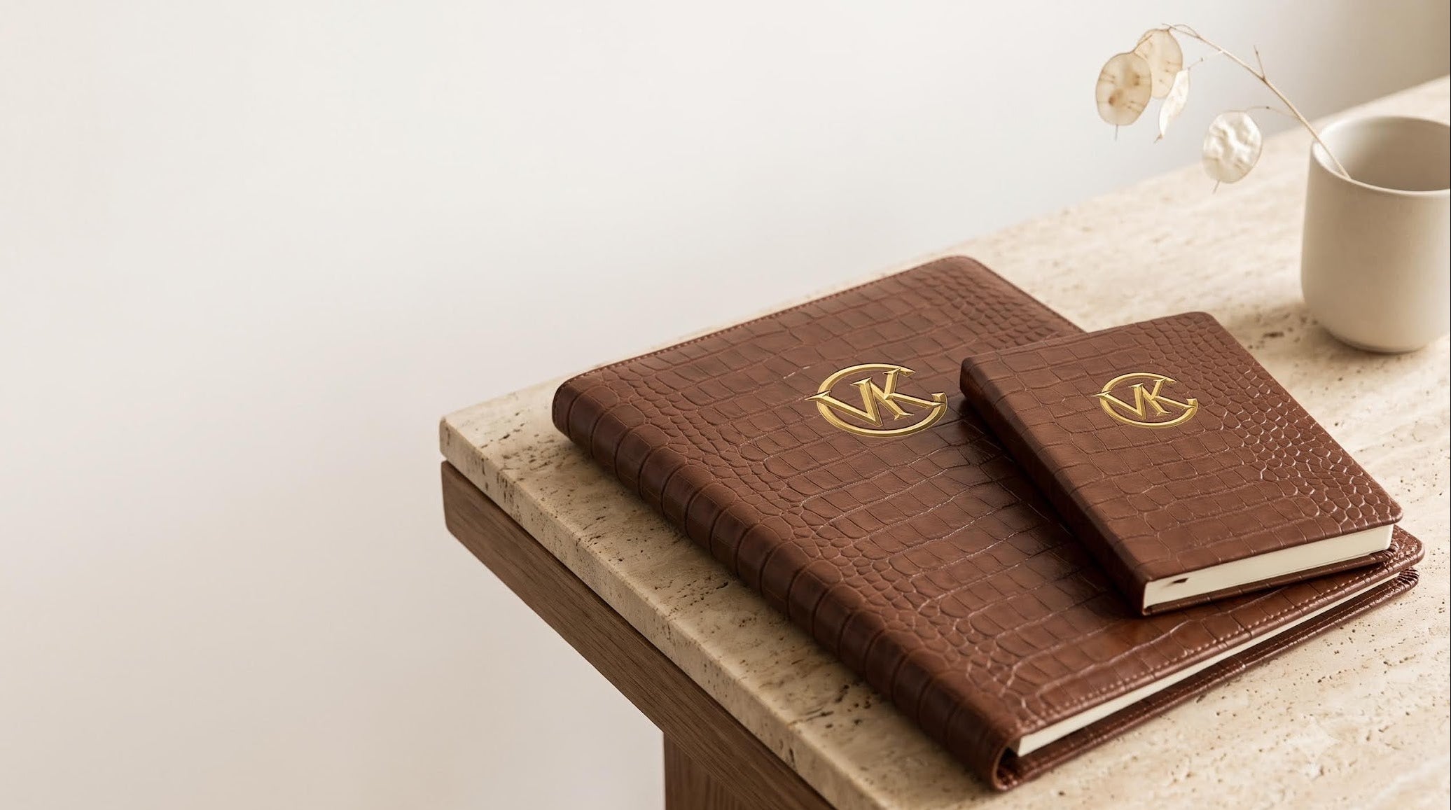 Two brown embossed leather notebooks with gold monogram covers resting on a light stone tabletop next to a white ceramic mug.